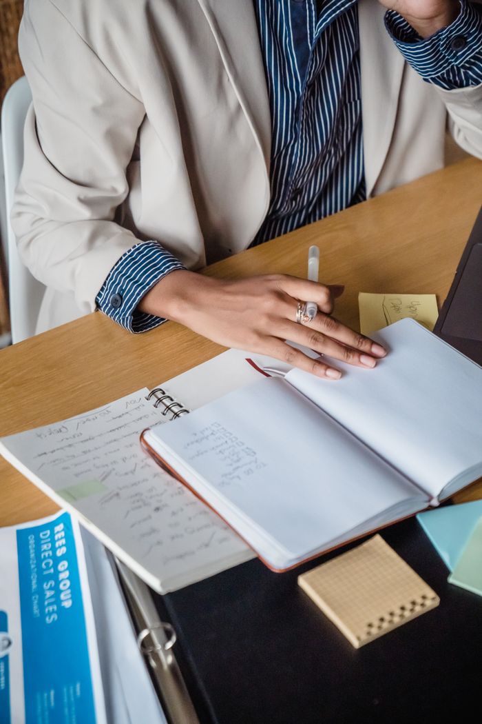 Woman writing in a notebook at a desk with papers and sticky notes, highlighting office productivity.